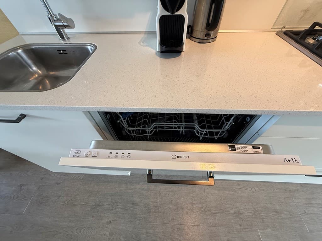 Uitzicht op een moderne keuken met een open vaatwasser, spoelbak en koffiezetapparaat op een lichtgekleurde countertop.