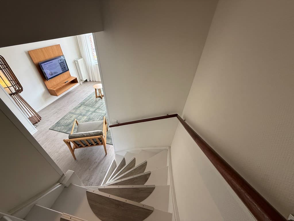 Uitzicht op een trap met witte treden en houten leuning naar een woonkamer met televisie en meubel in de verte
