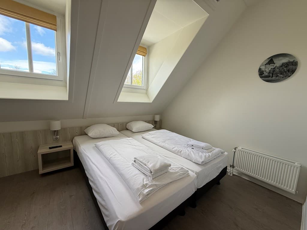 Slaapkamer met twee afzonderlijke bedden, kleine nachtkastjes en een radiator onder een schuin plafond met grote ramen