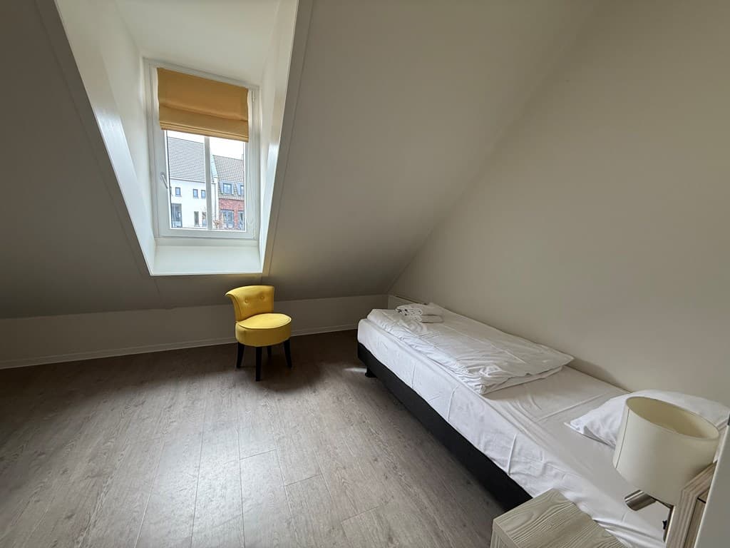 Slaapkamer met schuin dak, eenpersoonsbed en een gele stoel bij het raam met uitzicht op de buitenruimte.