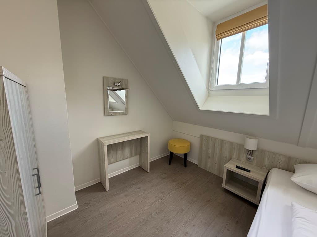 Slaapkamer met een schuin dakraam, een bed, een bijzettafel en een spiegel boven een houten kast