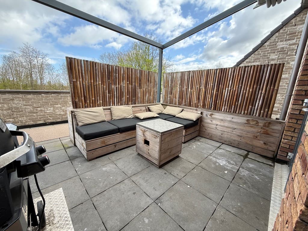 Terras met een loungebank van houten banken en kussens, een lage tafel en een barbecue in een houten omheining