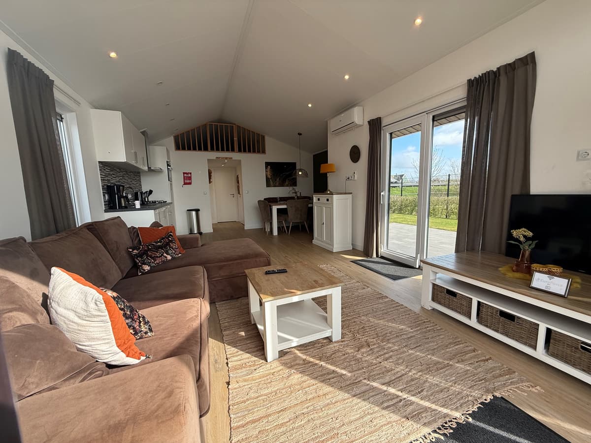 Woonkamer met een bruine bank, wit tv-meubel en openslaande deuren naar een terras met uitzicht op de natuur