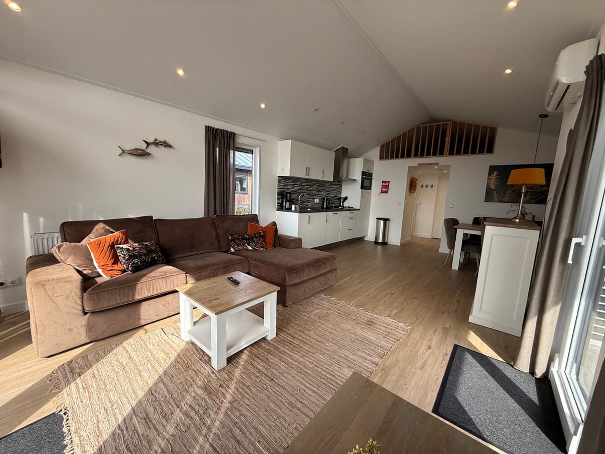 Lichte woonkamer met een grote bank, eettafel en open keuken met moderne apparatuur en houten vloeren.