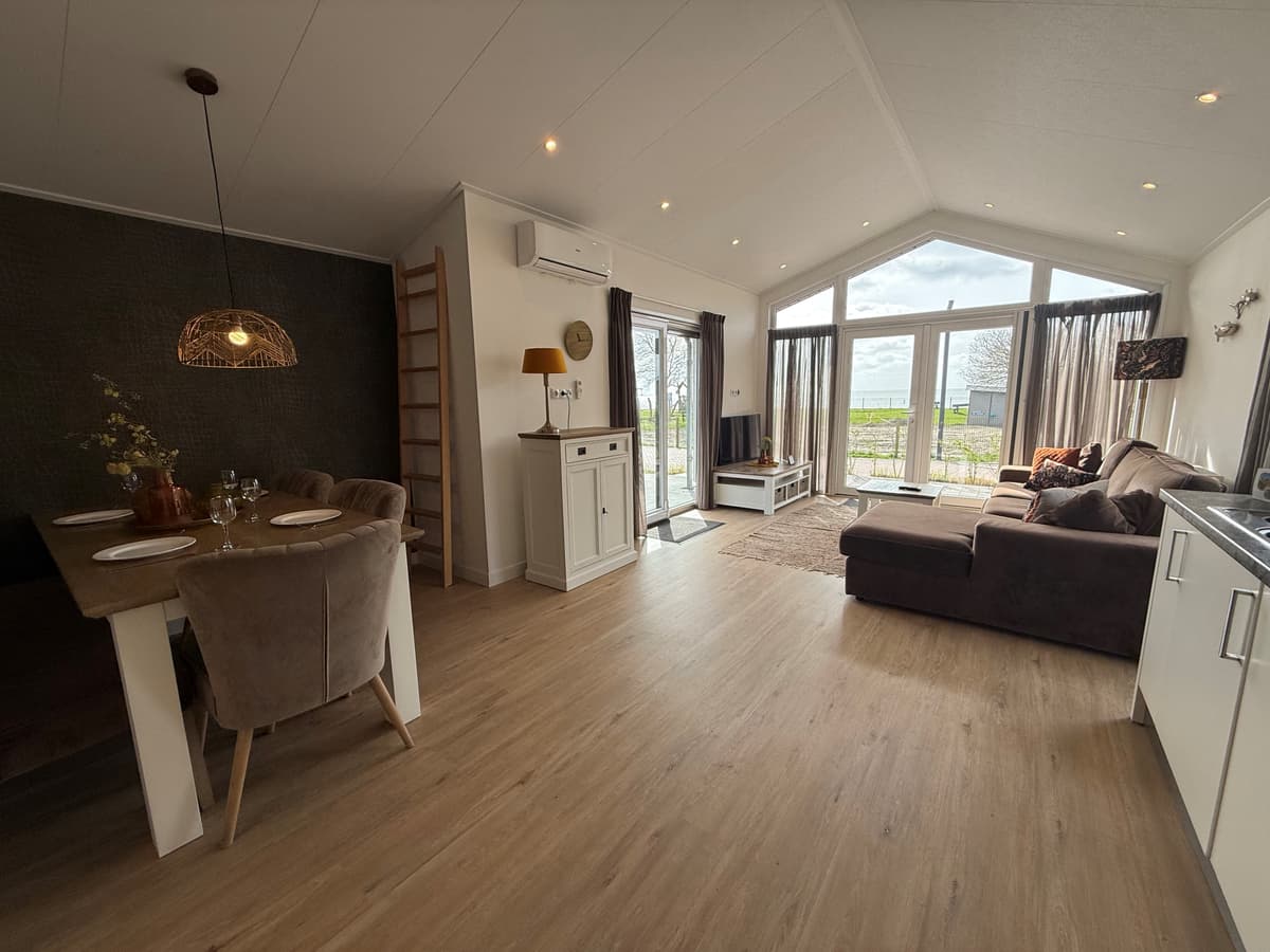 Licht interieur van een vakantiewoning met zithoek, eettafel en grote ramen met uitzicht op het buitengebied.