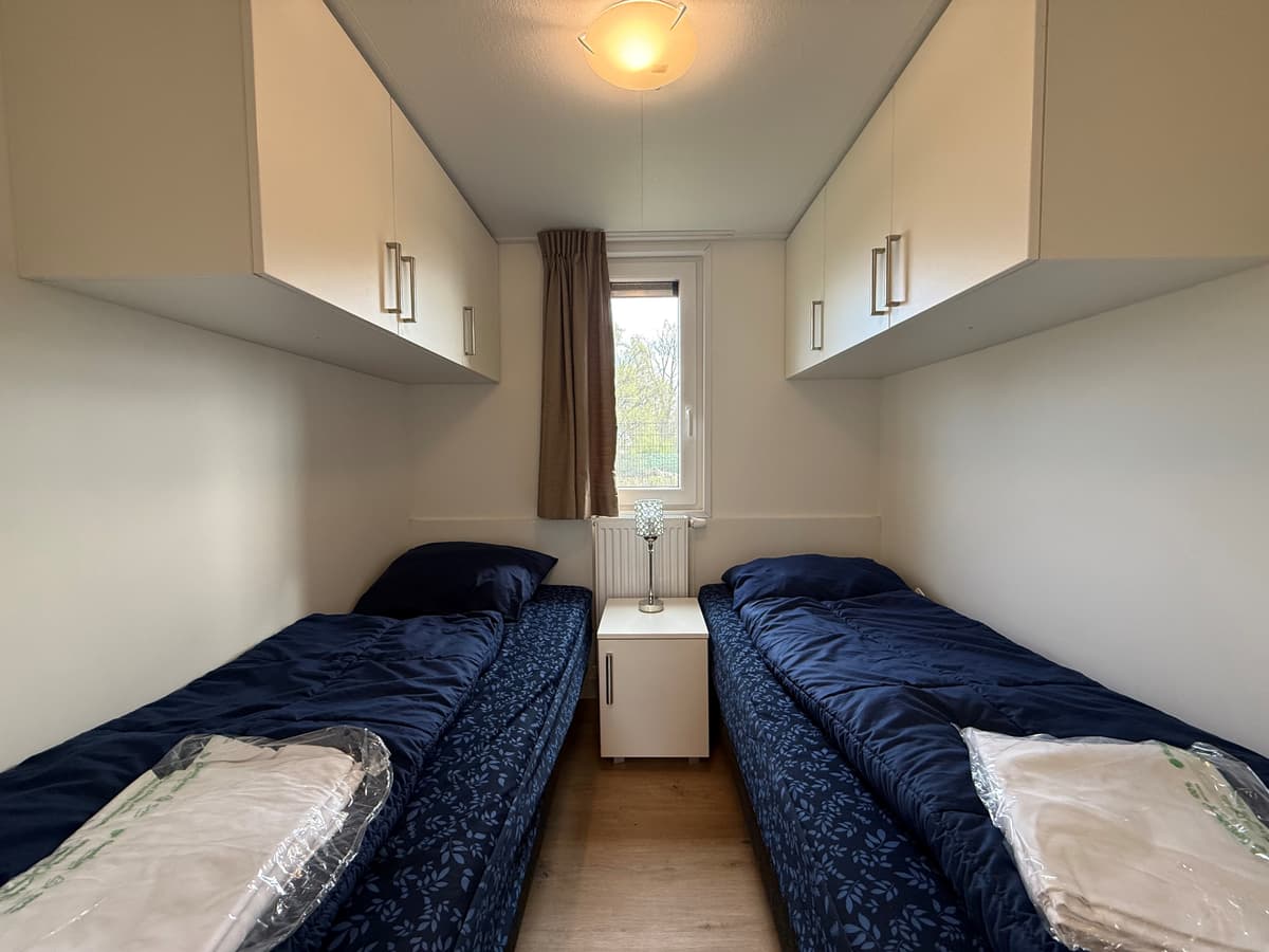 Slaapkamer met twee eenpersoonsbedden, nachtkastje en kastjes boven de bedden. Raam met gordijn zichtbaar.