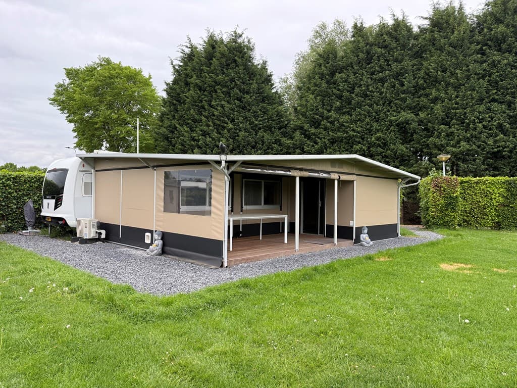 Tuin met een comfortabele caravan, een overkapping en een aangelegd terras, omringd door gras en heggen.