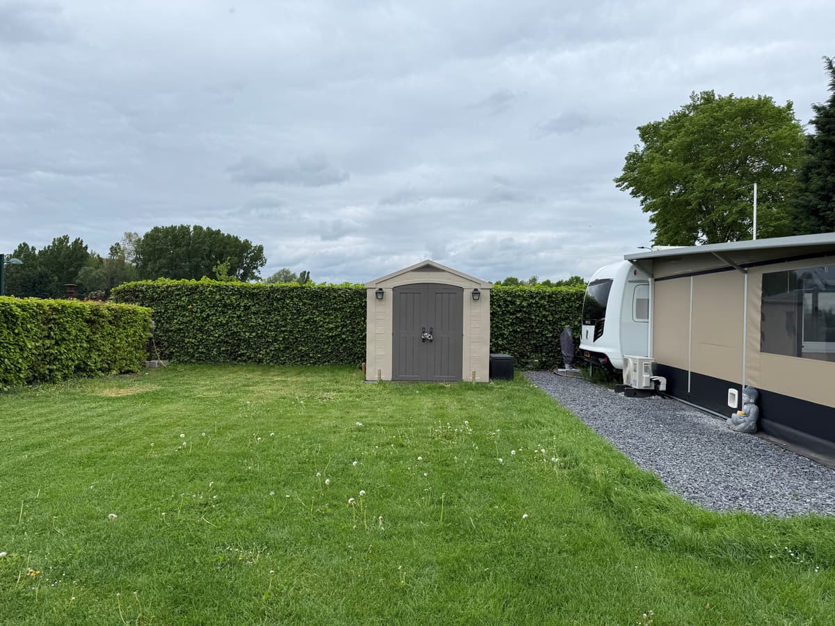 Tuin met gras, een schuur en een camper, omringd door groene heggen en een gravelpad.