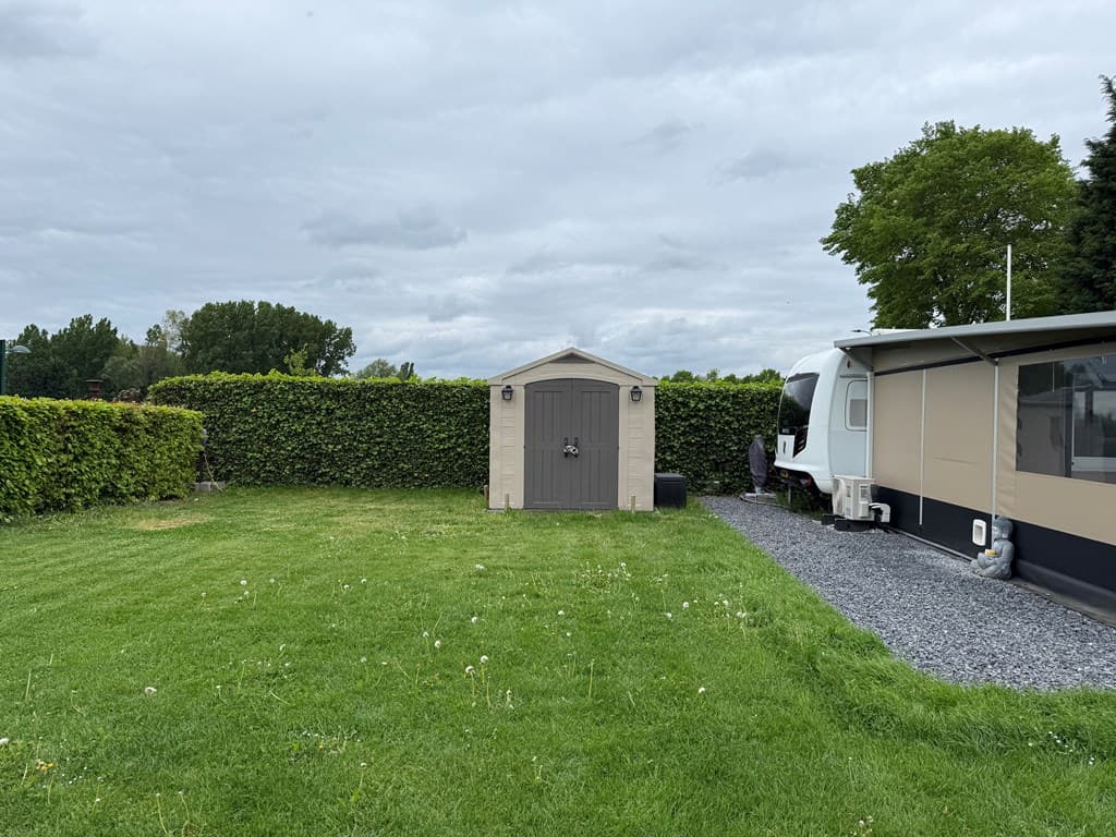 Tuin met gras, een schuur en een camper, omringd door groene heggen en een gravelpad.