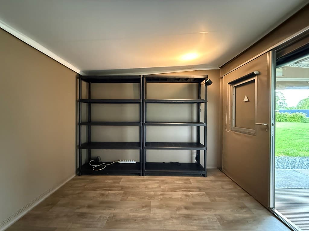 Ruime kamer met een houten vloer, zwarte open rekken en een deur naar de tuin met zicht op groen.