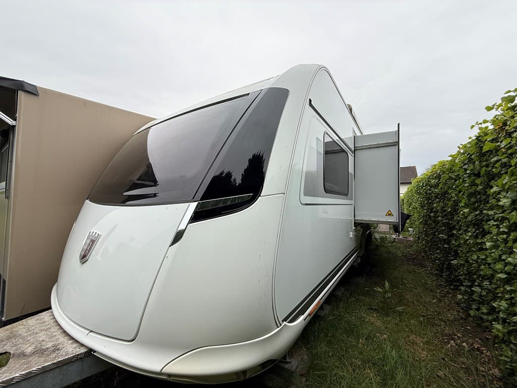 Rechthoekige caravan met grote ramen, geparkeerd naast een haag en een ander gebouw, op een grasachtige ondergrond.