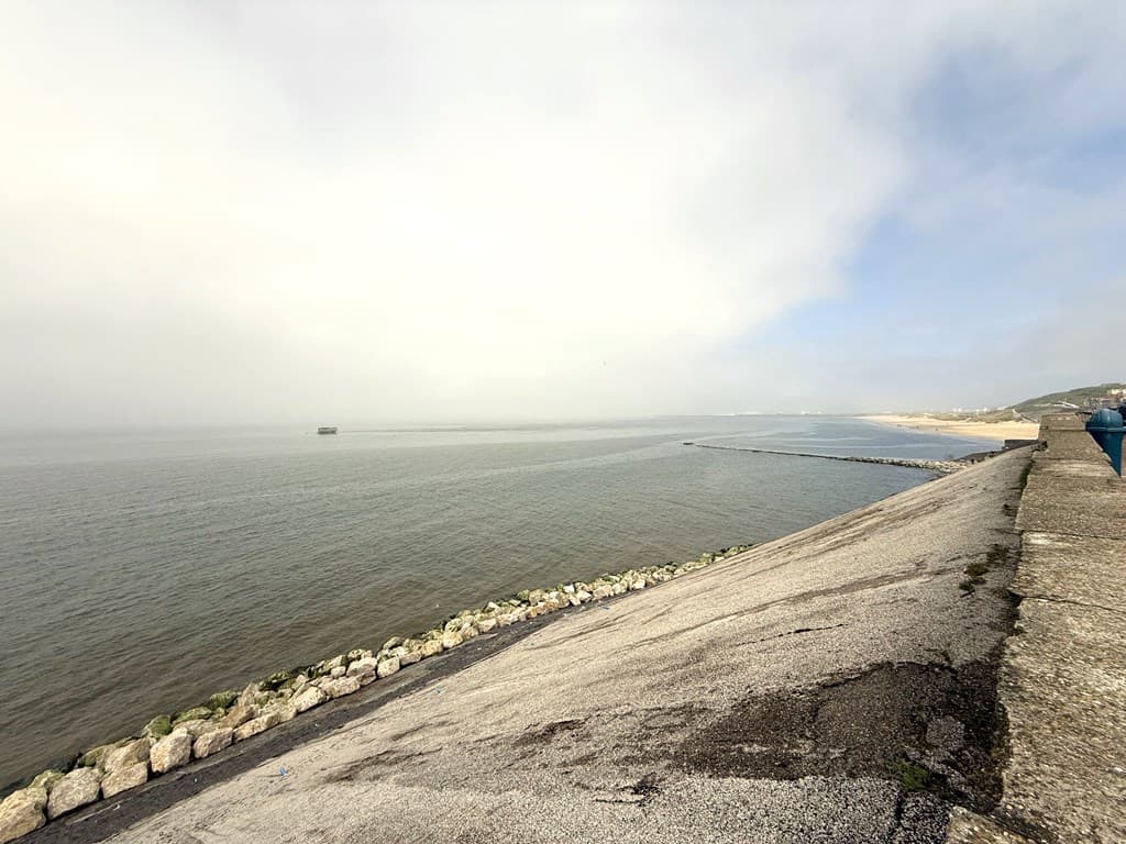Uitzicht over het water met een kiezelstrand en een betonnen oever, omgeven door mistige lucht en schepen in de verte