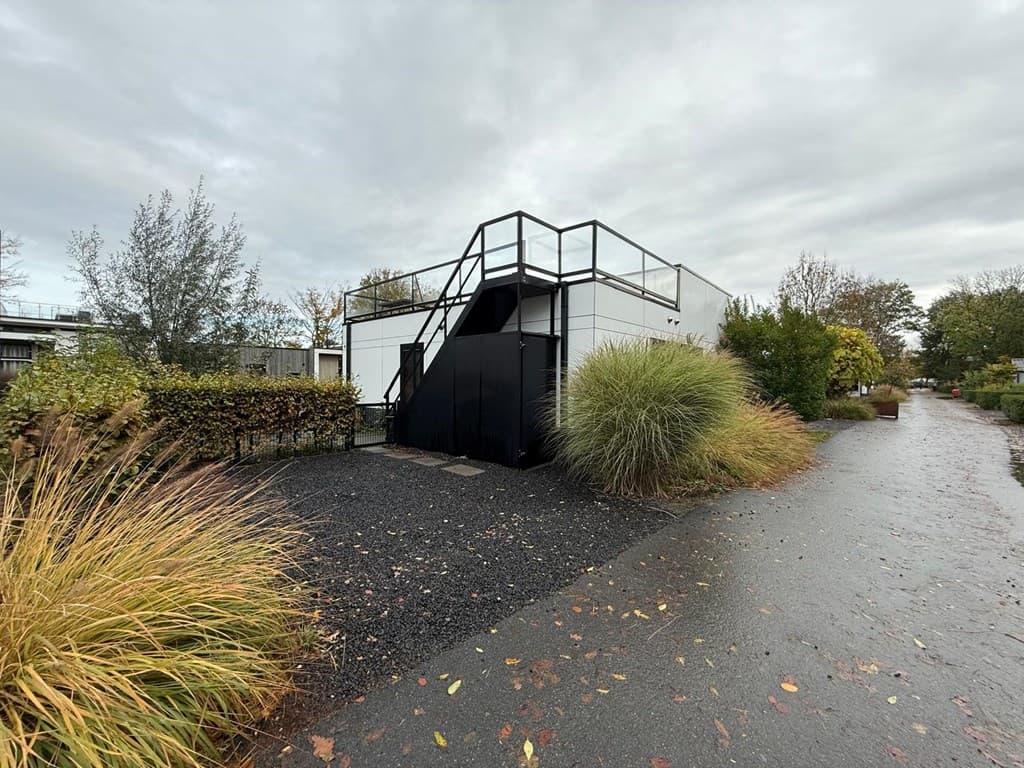 Moderne vakantiewoning met zwarte accenten en een trap, omgeven door groene hagen en een betegeld pad.