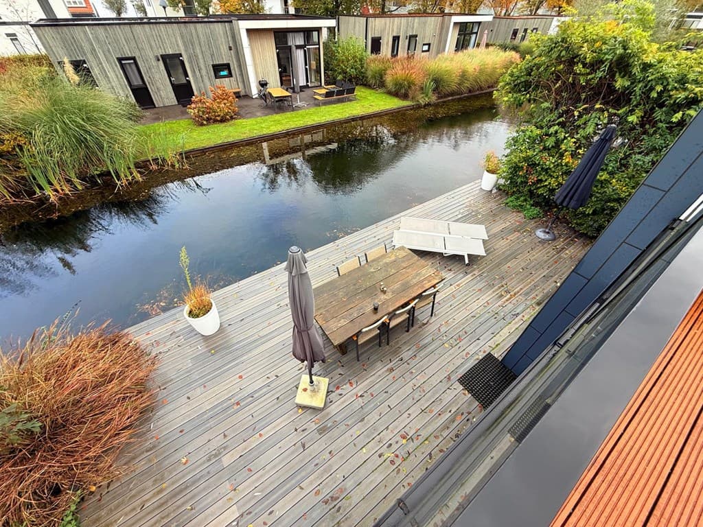 Terras aan het water met een houten tafel en stoelen, omringd door groene planten en watervlakte.