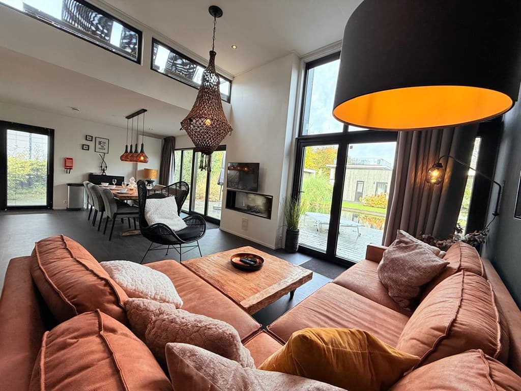 Knusse woonkamer met oranjerode bank, moderne verlichting en uitzicht op het terras met eetruimte en natuur.