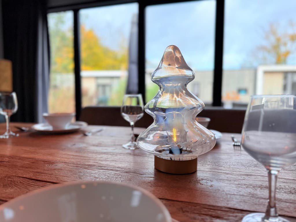 Tafel met glazen kerstboomlamp en servies met wijnglazen bij grote ramen met uitzicht op buiten.