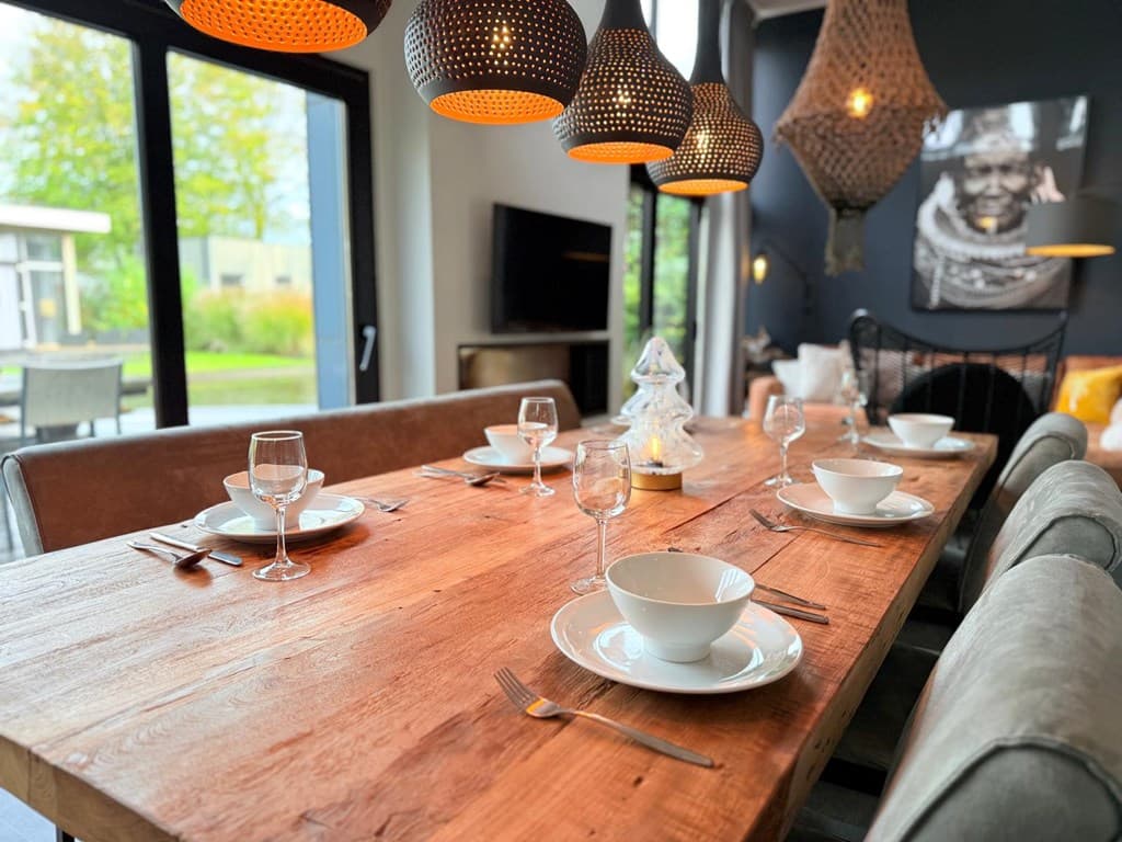 Eettafel met borden en glazen, moderne inrichting, grote ramen met uitzicht op de tuin en sfeervolle verlichting.