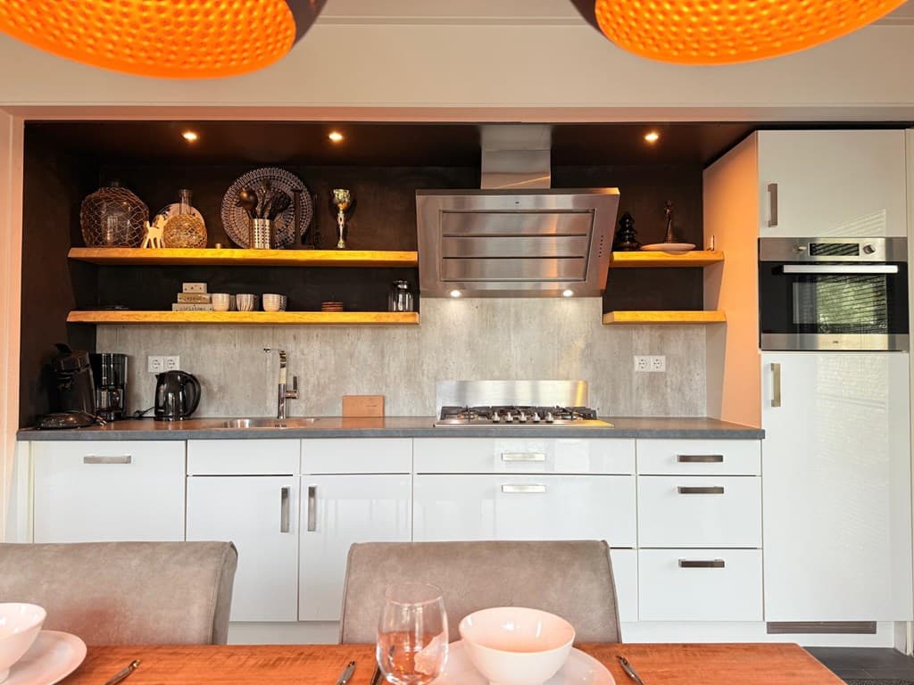 Moderne open keuken met witte kasten, apparatuur en houten planken boven het aanrecht. Eetruimte zichtbaar op de voorgrond.