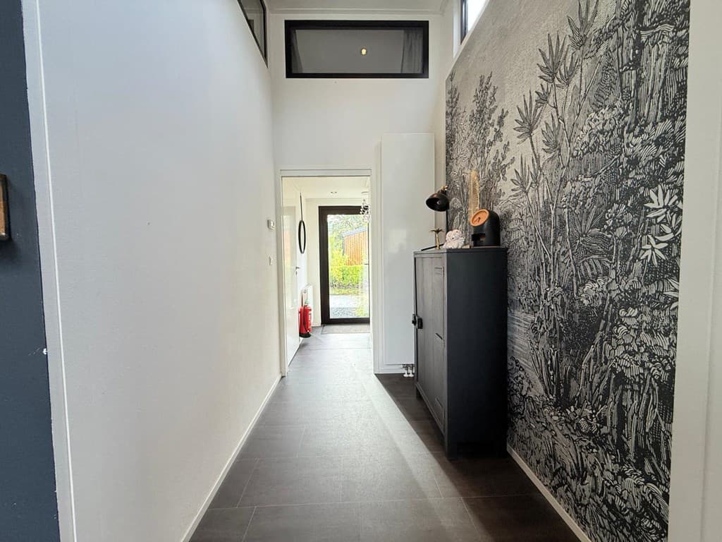 Lange, smalle hal met lichte muren en een wanddecoratie van planten, en toegang tot de voordeur aan het einde.