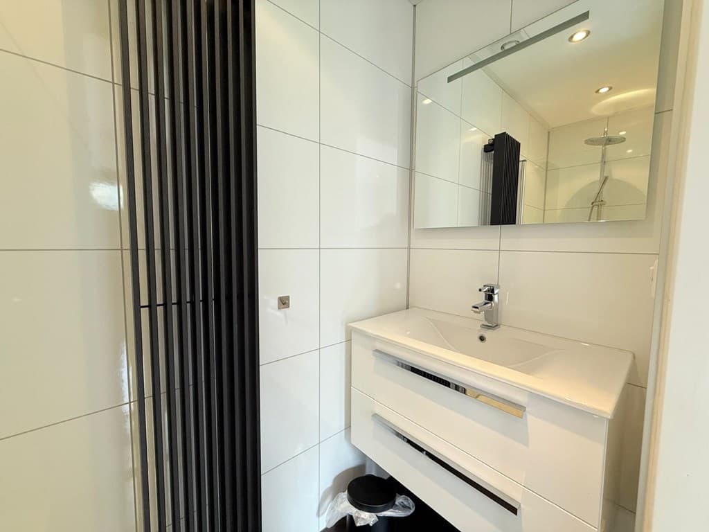 Moderne badkamer met witte wandtegels, inbouwverlichting, wastafel met spiegel en een douchecabine.