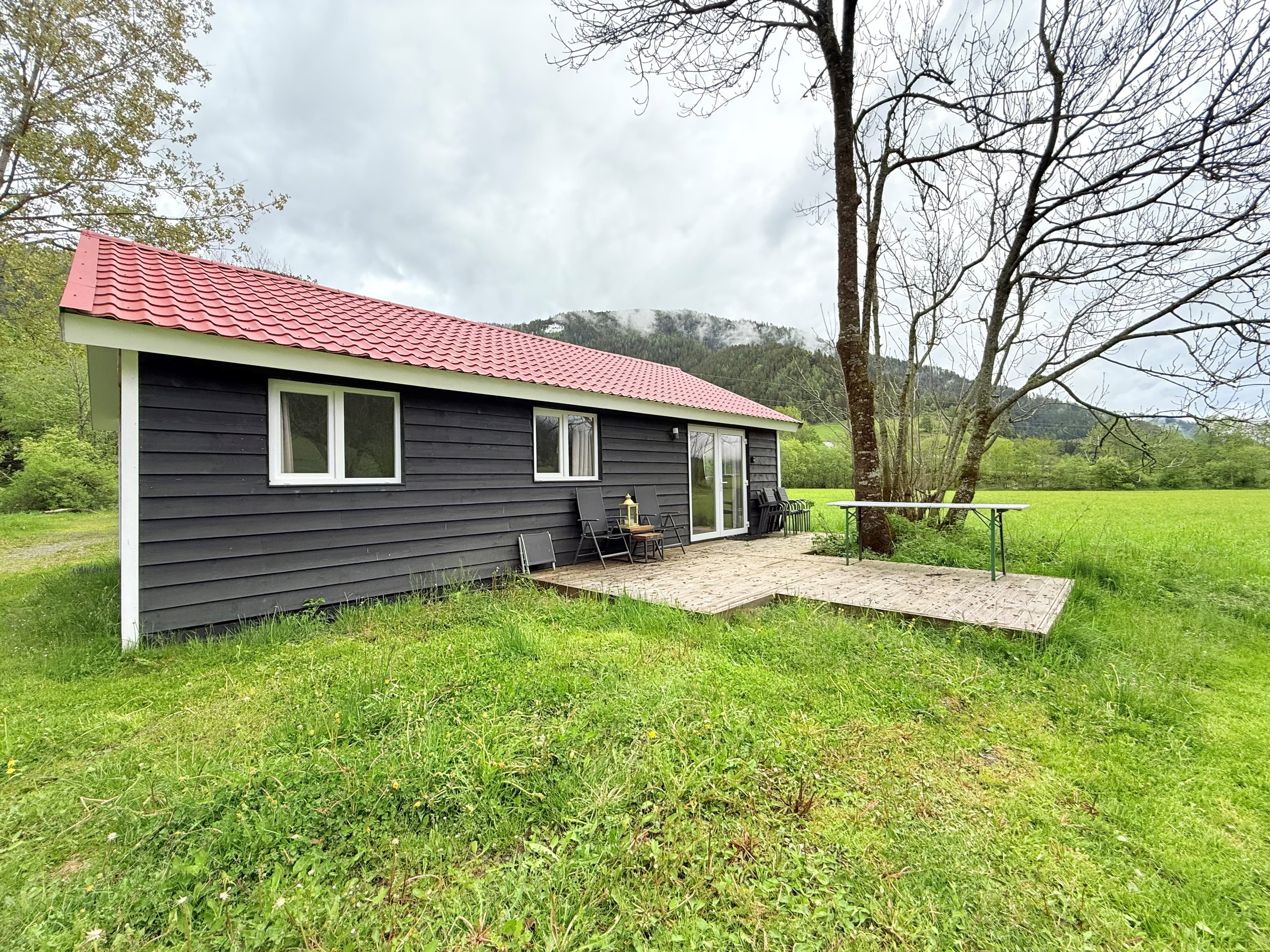 Zwart houten huis met rode dak, terras met tafeltje en stoelen, omgeven door groen gras en bomen met bergen op de achtergrond