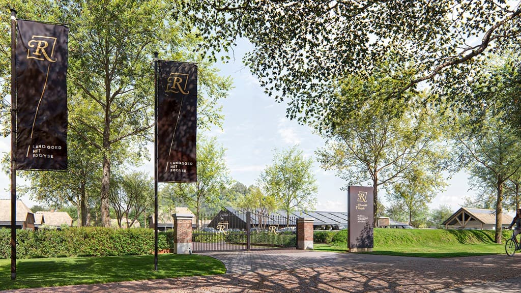 Ingang van het terrein met vlaggen en een toegangspoort omgeven door bomen en groen.