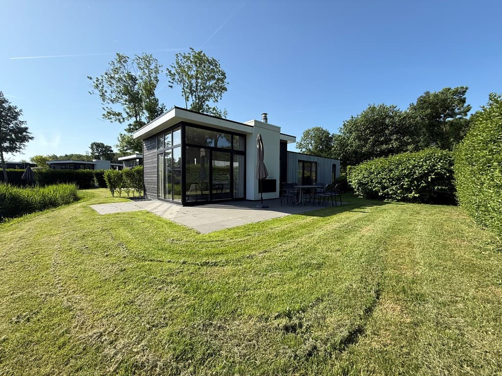 Moderne vakantiewoning omgeven door groen met een betegeld terras en grote ramen tegenover het grasveld.