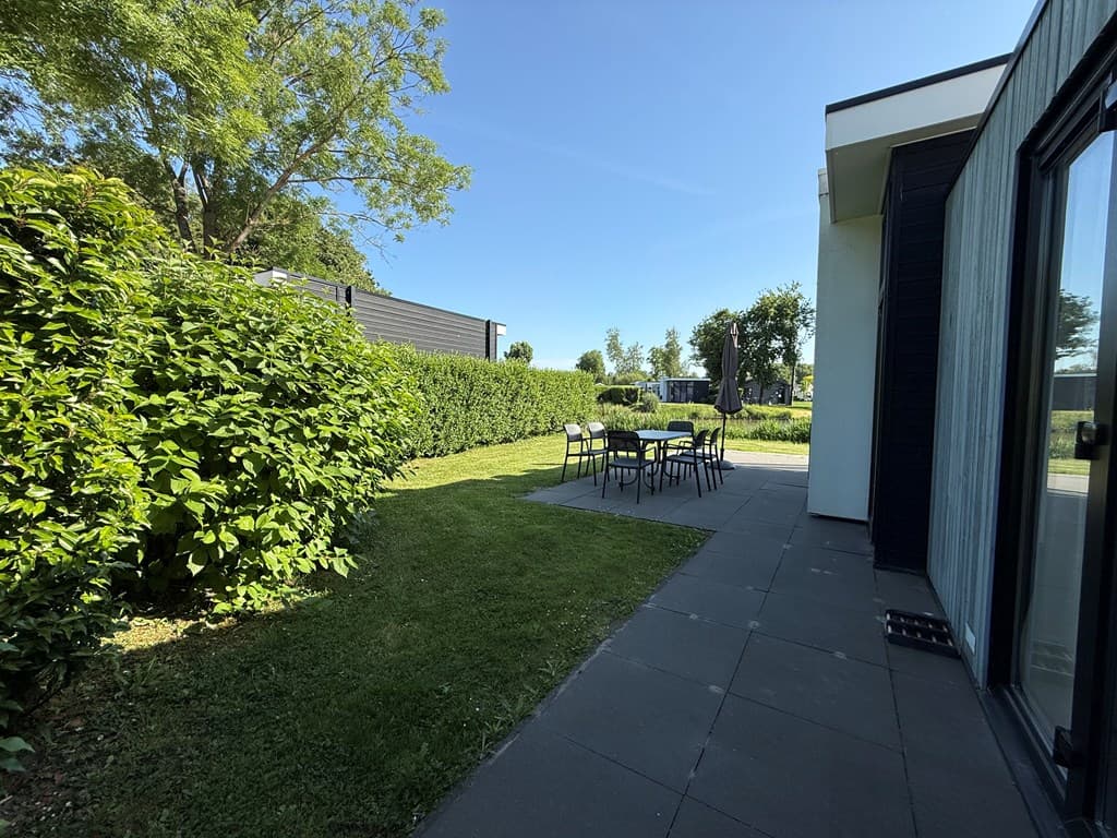 Tuin met grasveld en terras, omlijst door groene haag en struiken, met een tafel en stoelen op het terras