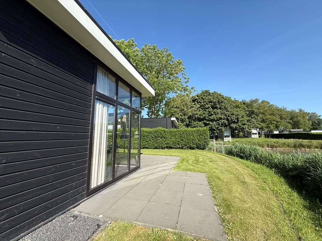 Zijaanzicht van een zwarte woning met grote ramen en betegeld terras, omringd door groen en grasvelden.