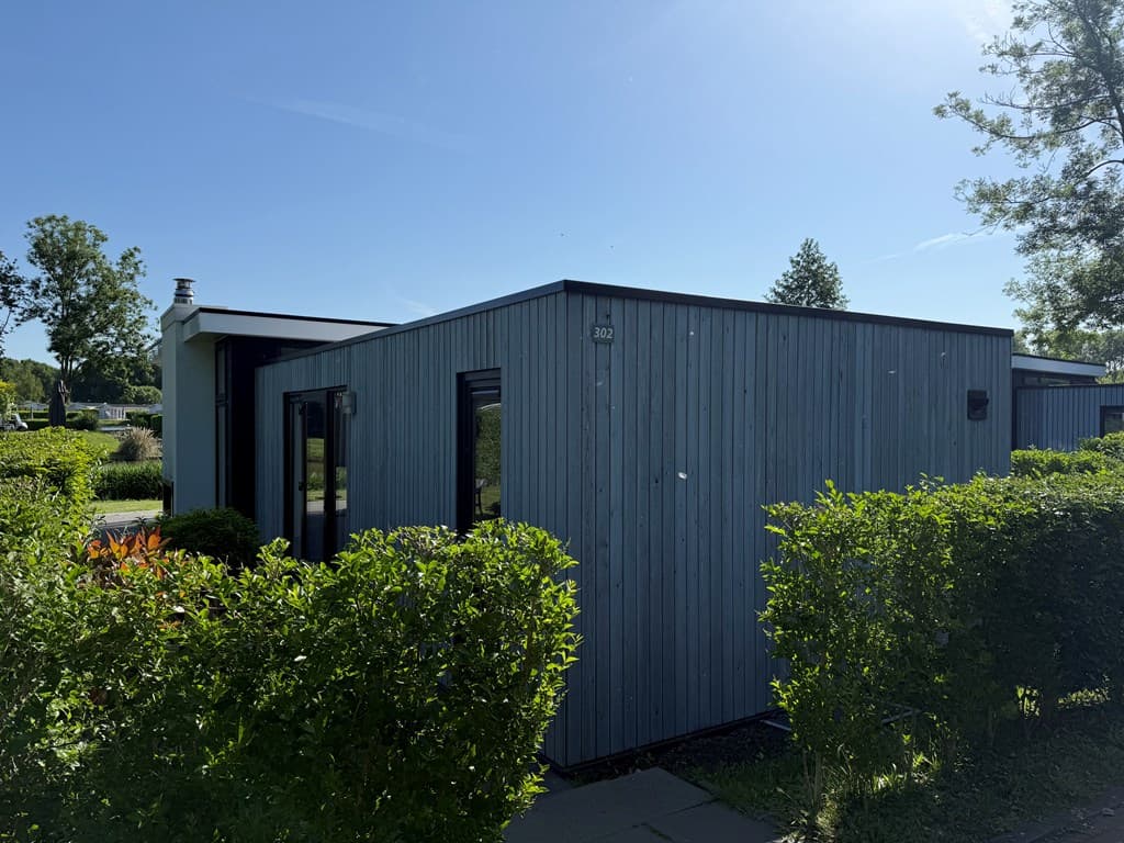Moderne vakantiewoning met houten gevel, omringd door groene struiken en zonnige blauwe lucht.