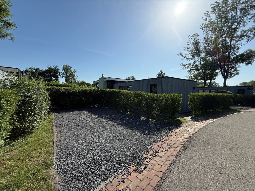 Grijs grind parkeerplaats omringd door heesterstruiken met bomen en woningen op de achtergrond onder een blauwe lucht