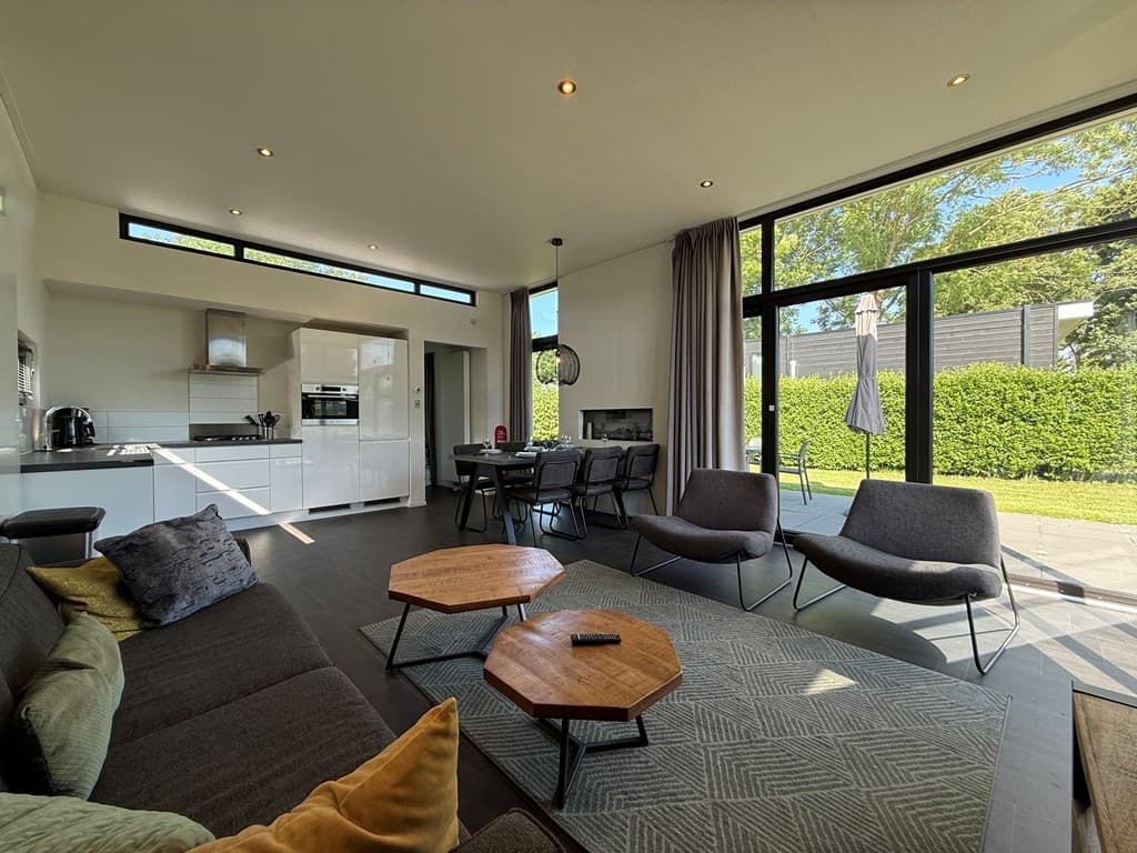 Moderne woonkamer met grote ramen, eetgedeelte en open keuken, zicht op de tuin met terras en meubilair.