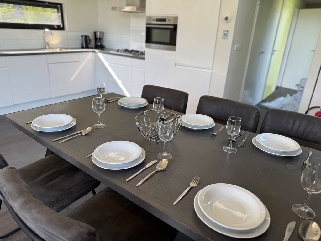 Eetgedeelte met een grote tafel, acht borden en glazen, moderne keuken op de achtergrond met apparatuur.
