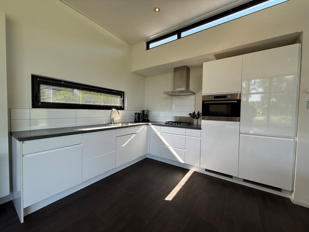 Moderne keuken met witte kastjes, gasfornuis, oven en zicht op buitenlicht via een raam.