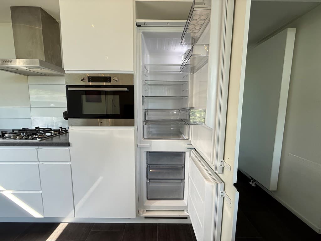 Keuken met een moderne koelkast, een kookplaat en een oven, alles in een strakke, witte afwerking.