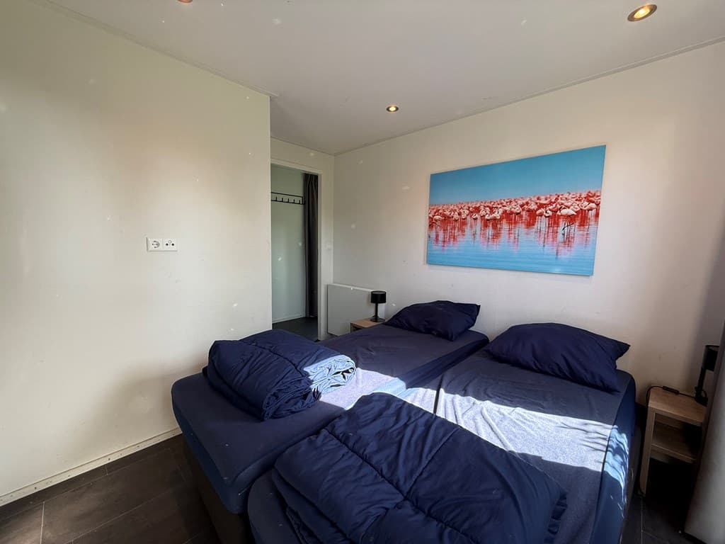 Licht ingerichte slaapkamer met twee losse bedden en een schilderij van een waterlichaam aan de muur.