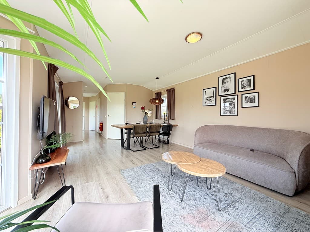 Woonkamer met bank, eettafel en fauteuil, foto's aan de muur en grote ramen met uitzicht op de groene omgeving
