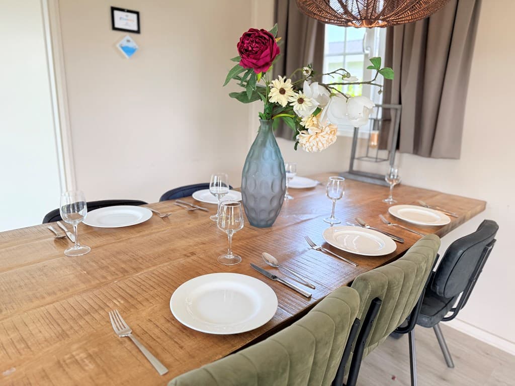 Eettafel met zes borden, glazen en bestek, een vaas met bloemen en een houten tafelblad in een lichte ruimte.