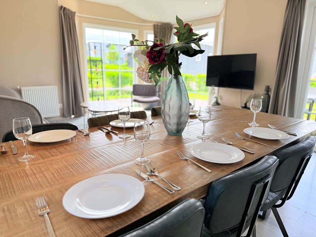 Eettafel met gedekte borden, glazen en een vaas met bloemen in een lichte woonkamer met grote ramen en uitzicht op groen.