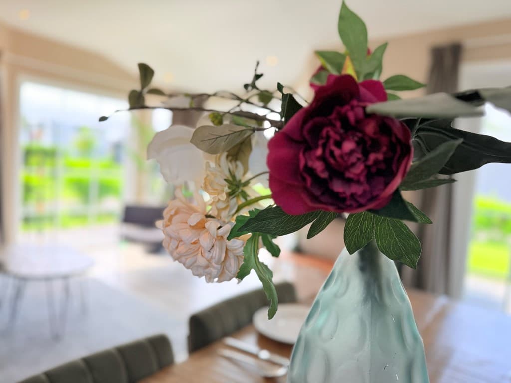 Tafel met een vaas met bloemen, op de achtergrond een lichte woonkamer met grote ramen en zicht op het tapeet.