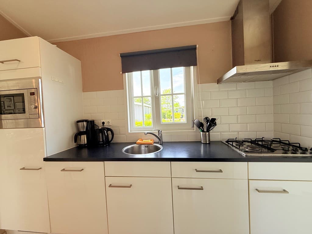 Keuken met witte kasten, spoelbak, gasfornuis en ramen met uitzicht op groen buiten.