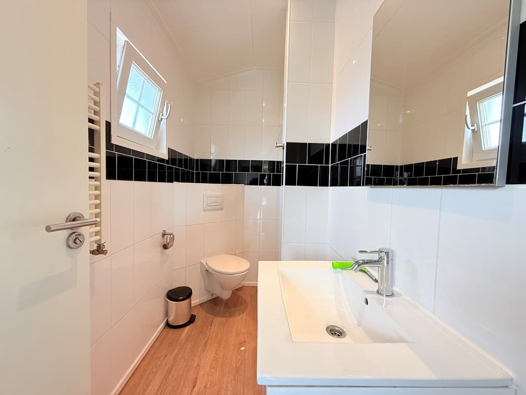 Badkamer met toilet, wastafel en spiegel, witte en zwarte wandtegels, raam aan de zijkant en vloerdetails.