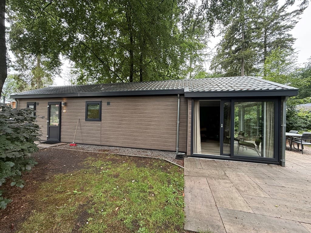 Moderne vakantiewoning met houten gevel, grote ramen en terras in een bosrijke omgeving.