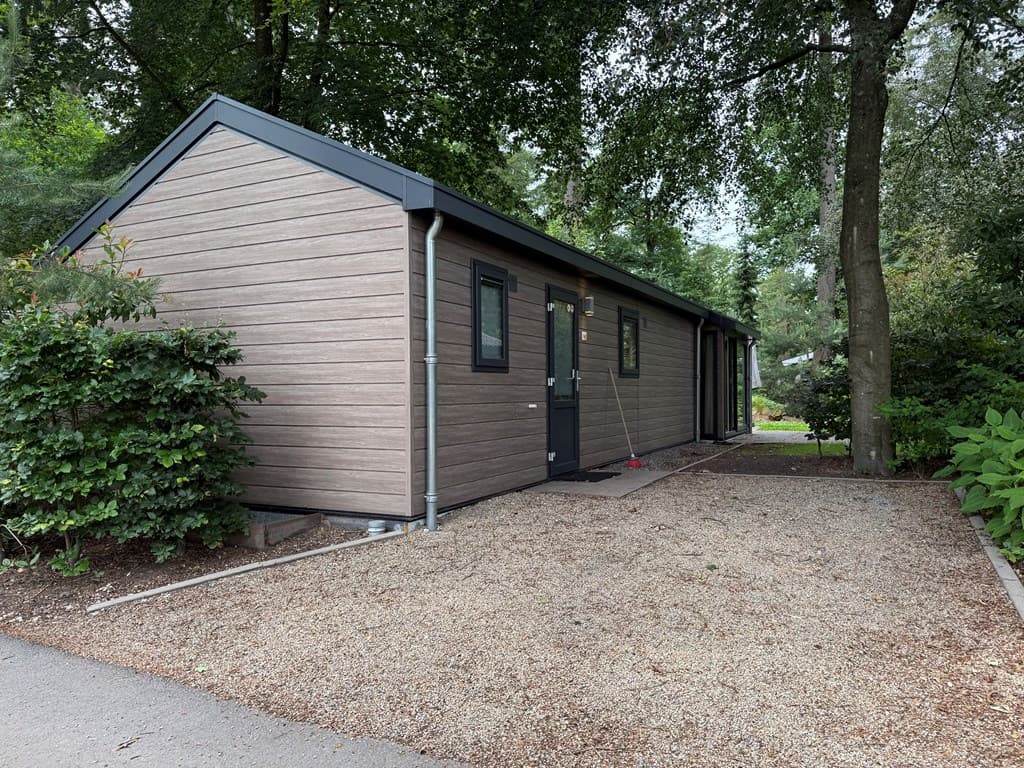 Vakantiehuis met houten buitenkant en een grindpad, omringd door bomen en groene planten in een rustige omgeving.