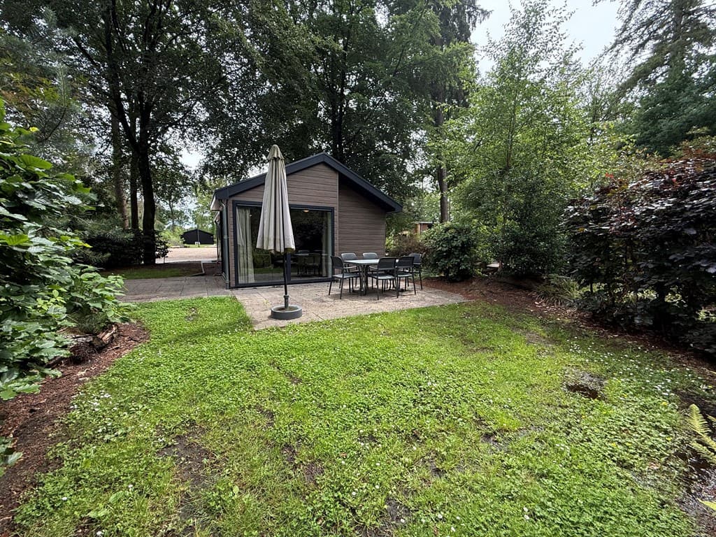 Buitenaanzicht van een vakantiewoning met terras en tuintafel onder een parasol, omringd door bomen en groene begroeiing