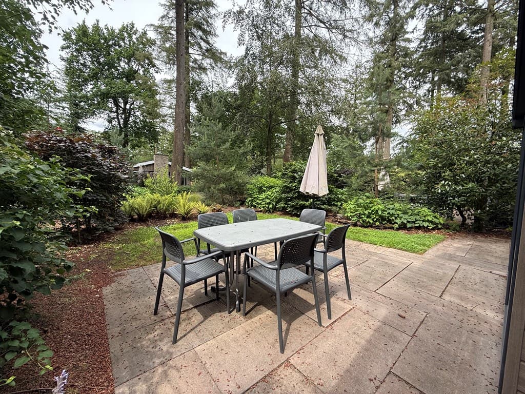 Tuin met een betegeld terras, een tafel met zes stoelen en een parasol, omringd door planten en bomen.