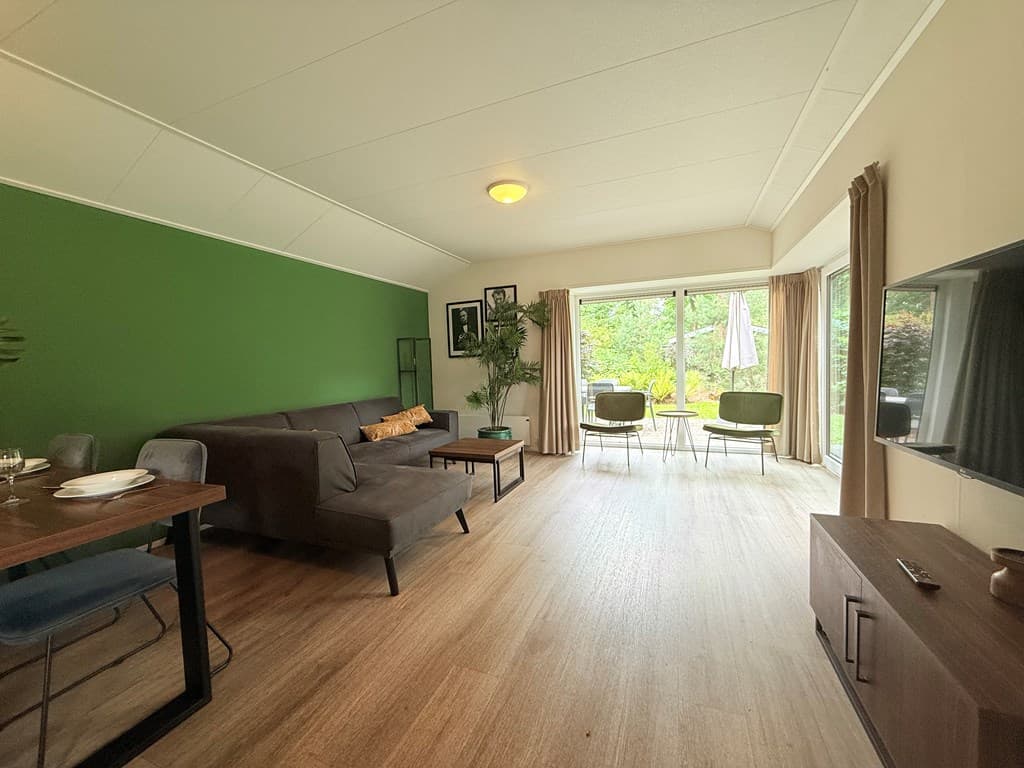 Ingerichte woonkamer met groene muur, zithoek, eettafel en toegang tot een terras met uitzicht op de tuin.