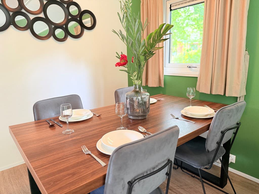 Eetkamer met een houten tafel, vier stoelen, servies en een vaas met bloemen bij een raam met gordijnen.