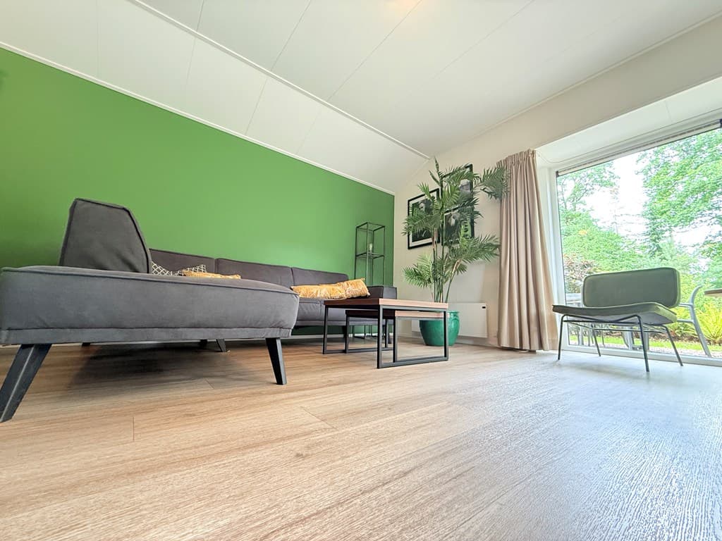 Zithoek met groen accentmuur, moderne meubels en veel lichtinval door grote raampartij met uitzicht op buiten.