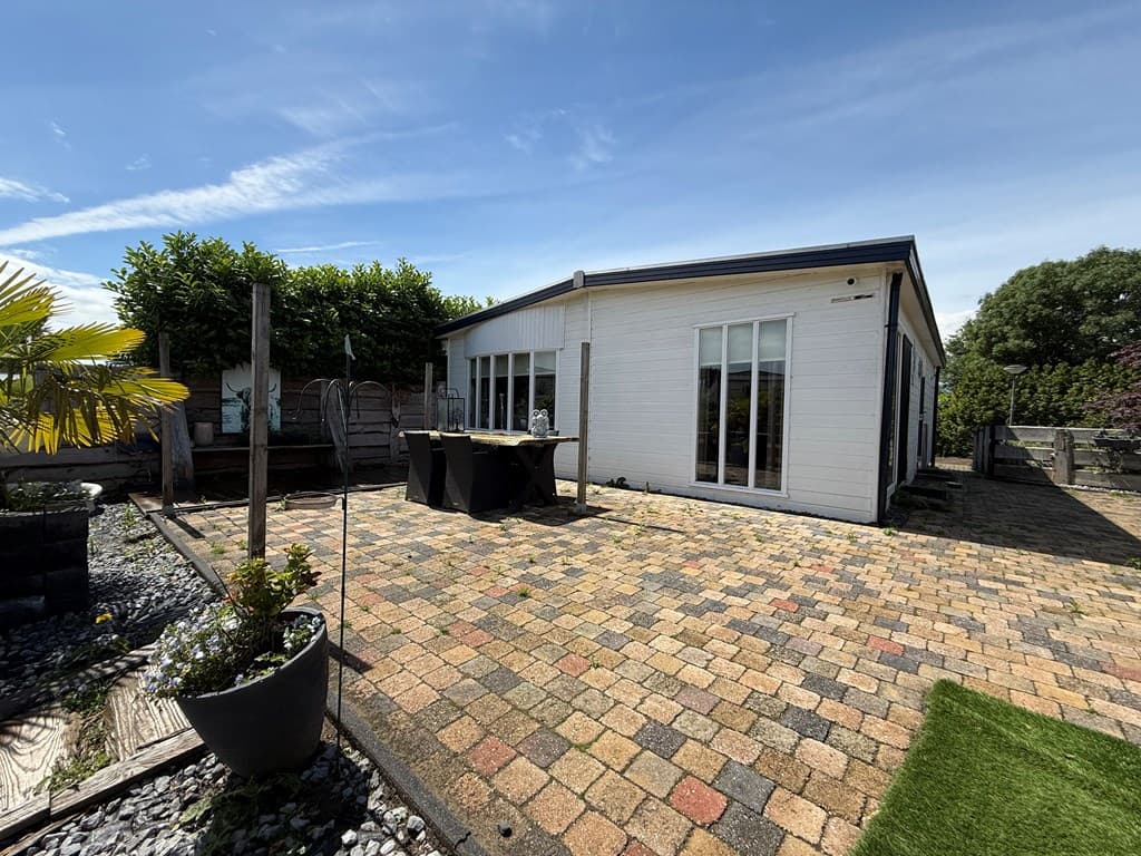 Woning met betegelde patio, planter met planten en een tafel met stoelen, omringd door groen en een schutting.
